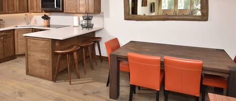 A kitchen with wooden cabinets, a stainless steel microwave, a sink, and a bar area with stools. There is a dining table with orange chairs and a large mirror above it.