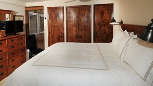 Un dormitorio con una cama grande, un armario de madera, un televisor sobre una cómoda y una ventana con persianas.