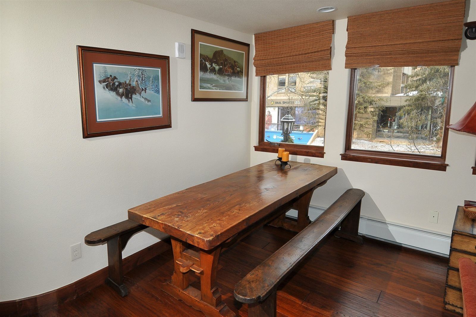 Una mesa de madera para comer con bancos, cuadros en la pared y vista a una piscina exterior.