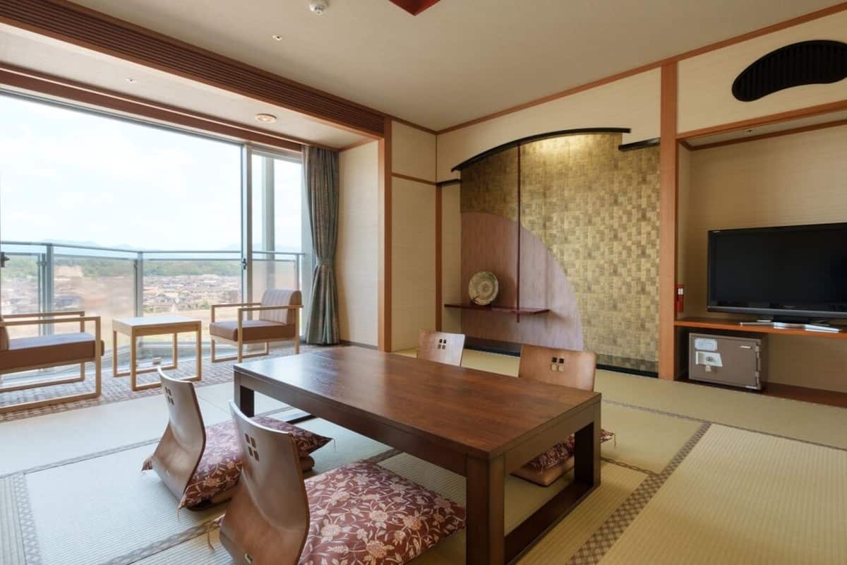 A hotel room with tatami flooring, a wooden dining table, chairs, a television, and a view of the landscape.