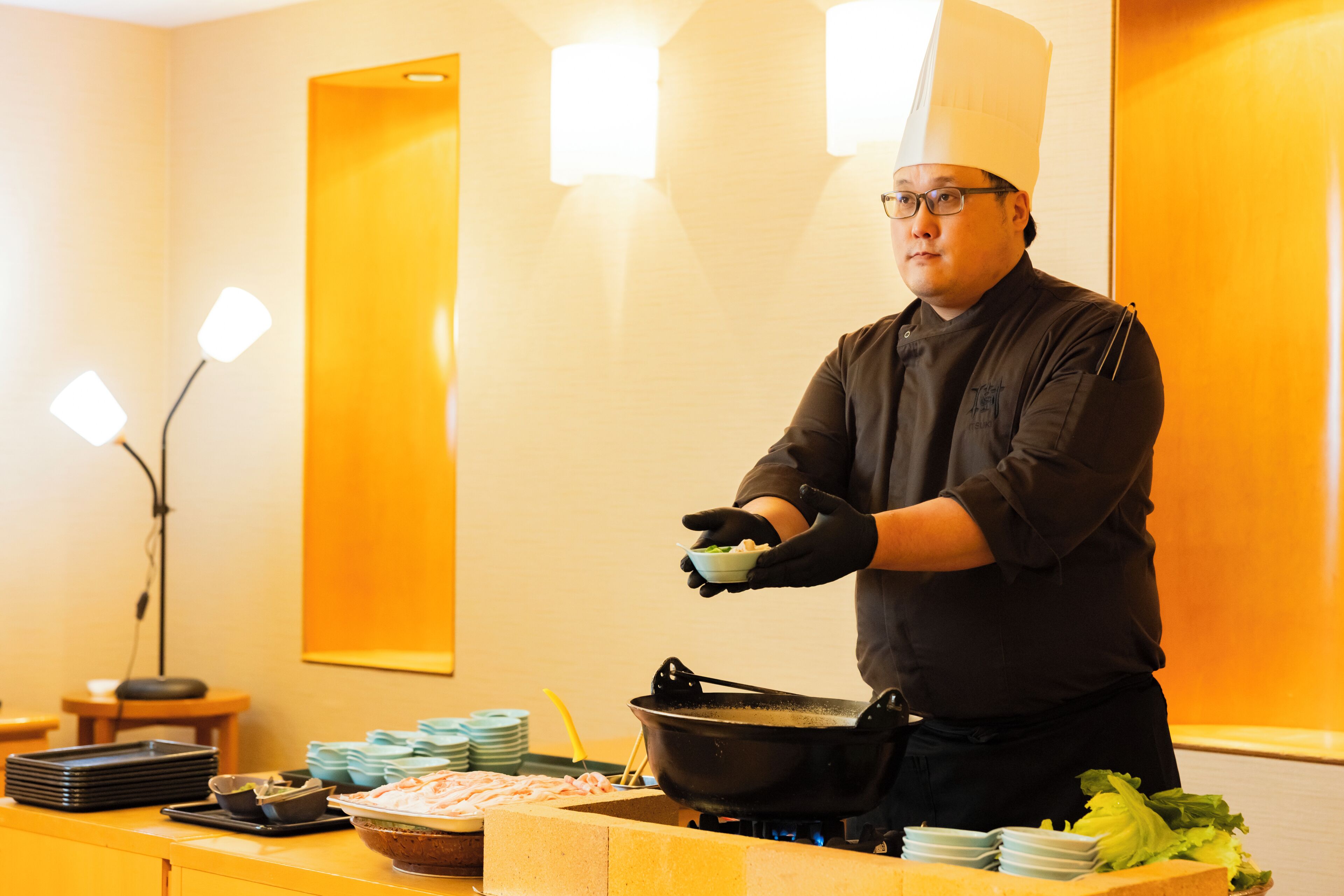 A chef in a professional kitchen setting with a stove, cooking utensils, and food preparation items.