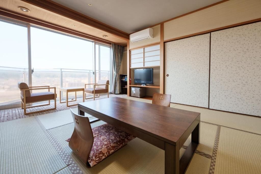 A hotel room with a large sliding glass door to a balcony, a wooden table, two chairs, a television, and traditional Japanese sliding doors.