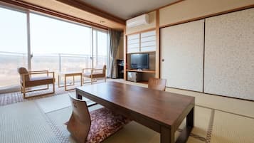 A hotel room with a large sliding glass door to a balcony, a wooden table, two chairs, a television, and traditional Japanese sliding doors.