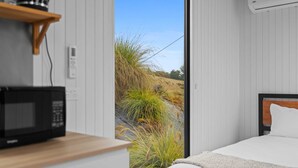 A bedroom with a bed, a wooden nightstand, a microwave, and a view of a grassy outdoor area through a window. - Bacardi Heights Waikaia by Tiny Away (Waikaia)
