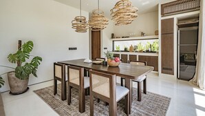 A modern dining area with a wooden table and chairs, a large potted plant, and a kitchen in the background. - Villa Bōld 01 At Bōld - Luxury Villas (Pecatu)