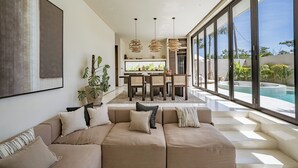 A modern living room with a beige sofa, a dining area with a table and chairs, and a large window overlooking a swimming pool. - Villa Bōld 01 At Bōld - Luxury Villas (Pecatu)