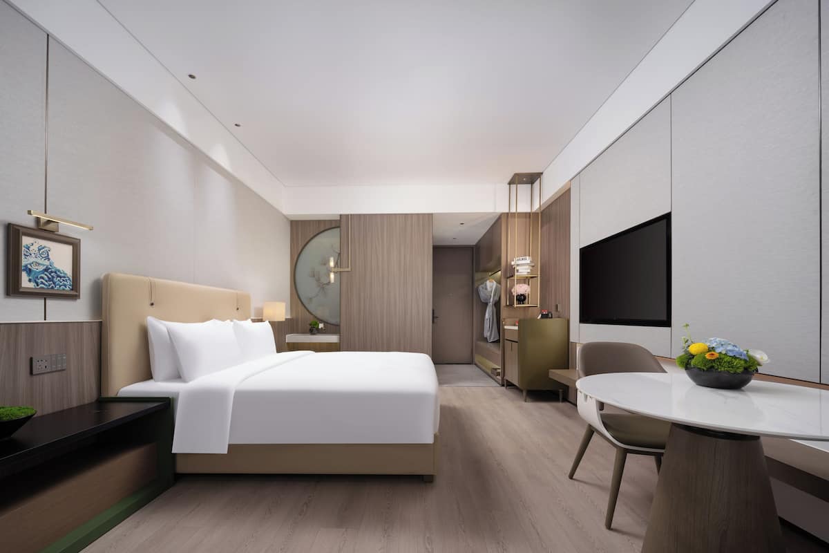 A modern hotel room with a bed, bedside table, TV, and a small dining area.