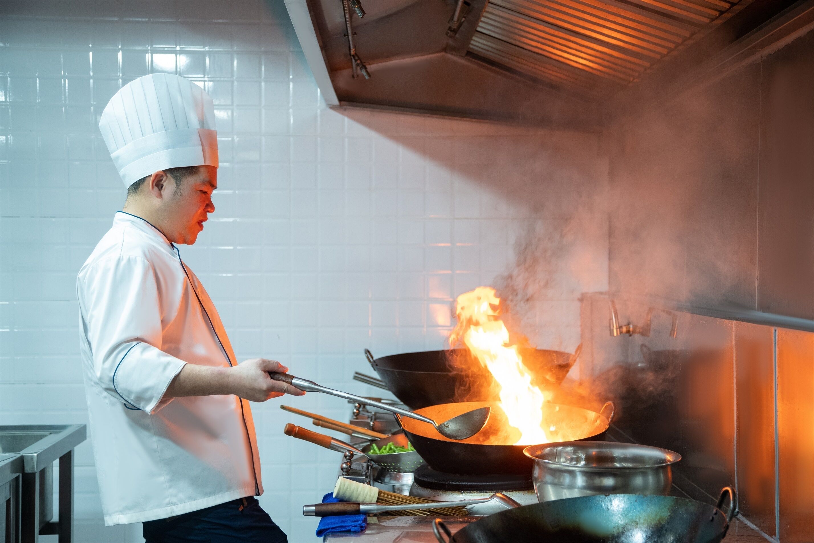 Un cocinero prepara comida en un wok sobre una hornilla, rodeado de utensilios de cocina y un salpicadero de azulejos.