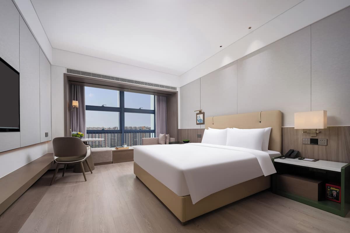 A modern hotel room with a large bed, a desk, a chair, and a view of the city through a large window.