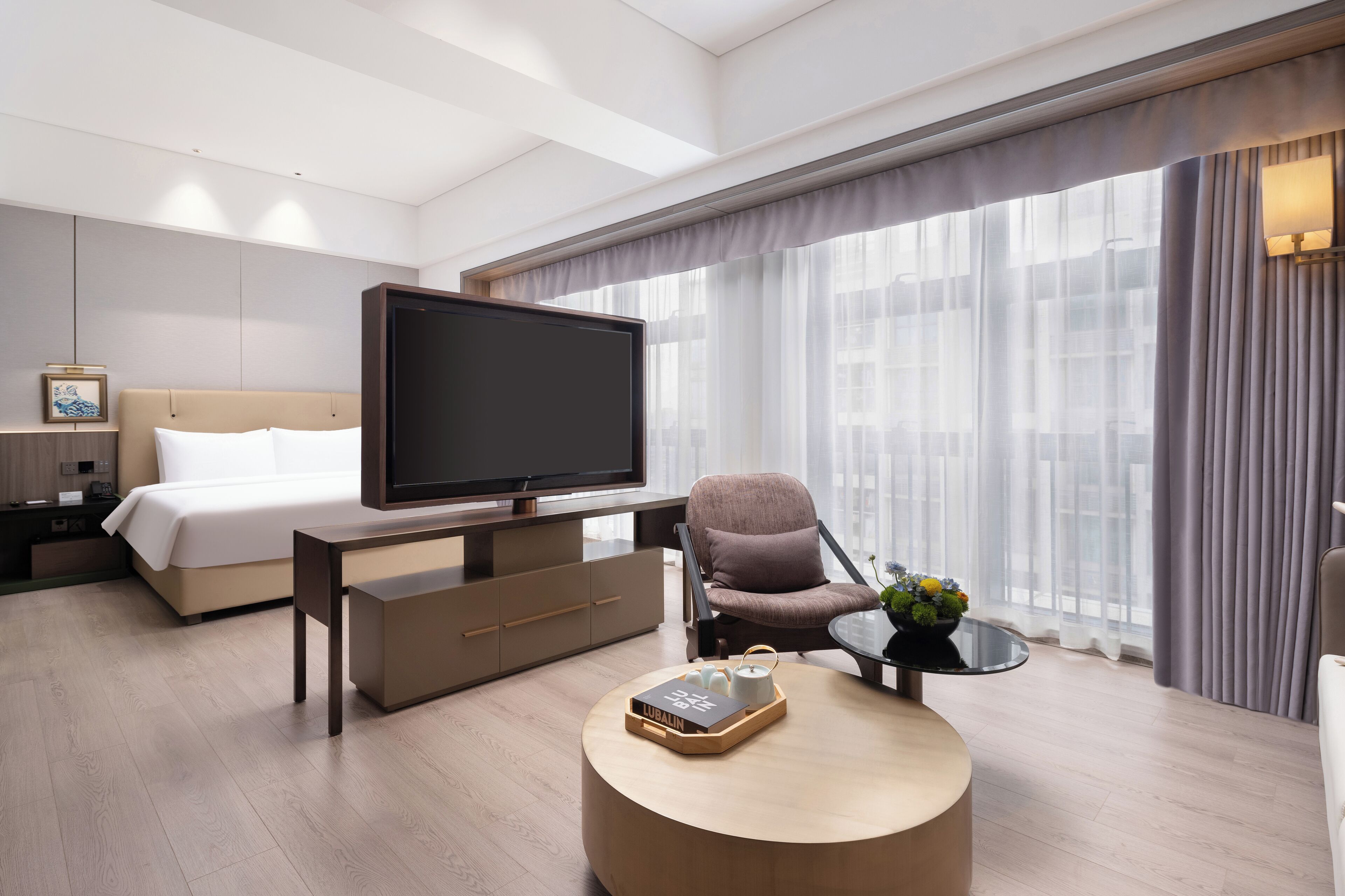Ein modernes Hotelzimmer mit einem großen Bett, einem Flachbildfernseher, einem runden Couchtisch mit Tablett und Blick auf die Stadt durch große Fenster.
