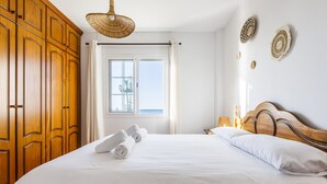 A neatly made bed with white linens and rolled towels, a wooden wardrobe, a window with a view, and a woven light fixture.