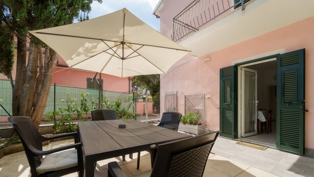 An outdoor patio with a table and chairs, a large umbrella, and a building entrance with green shutters. - APPARTAMENTO VIA DEL MARE 20 - VIA DEL MARE 20 (Marciana)