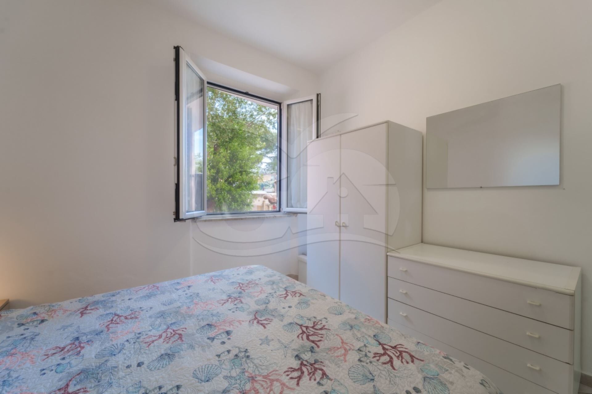 Een slaapkamer met een bed, een dressoir, een spiegel en een raam met uitzicht op bomen.