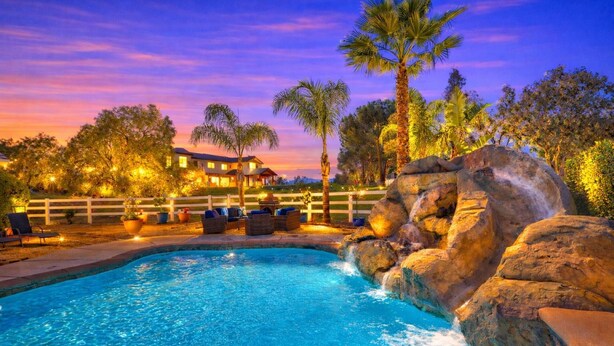 A swimming pool with a waterfall feature, surrounded by palm trees and a wooden fence. - Book Your Stay at Casa da Saudade | Pool, Slide, & Wine Country Views (Temecula)