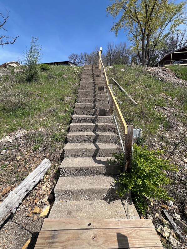 Un conjunto de escalones de piedra al aire libre que suben una colina, con una barandilla de madera a la derecha y una tabla de madera en la parte inferior.