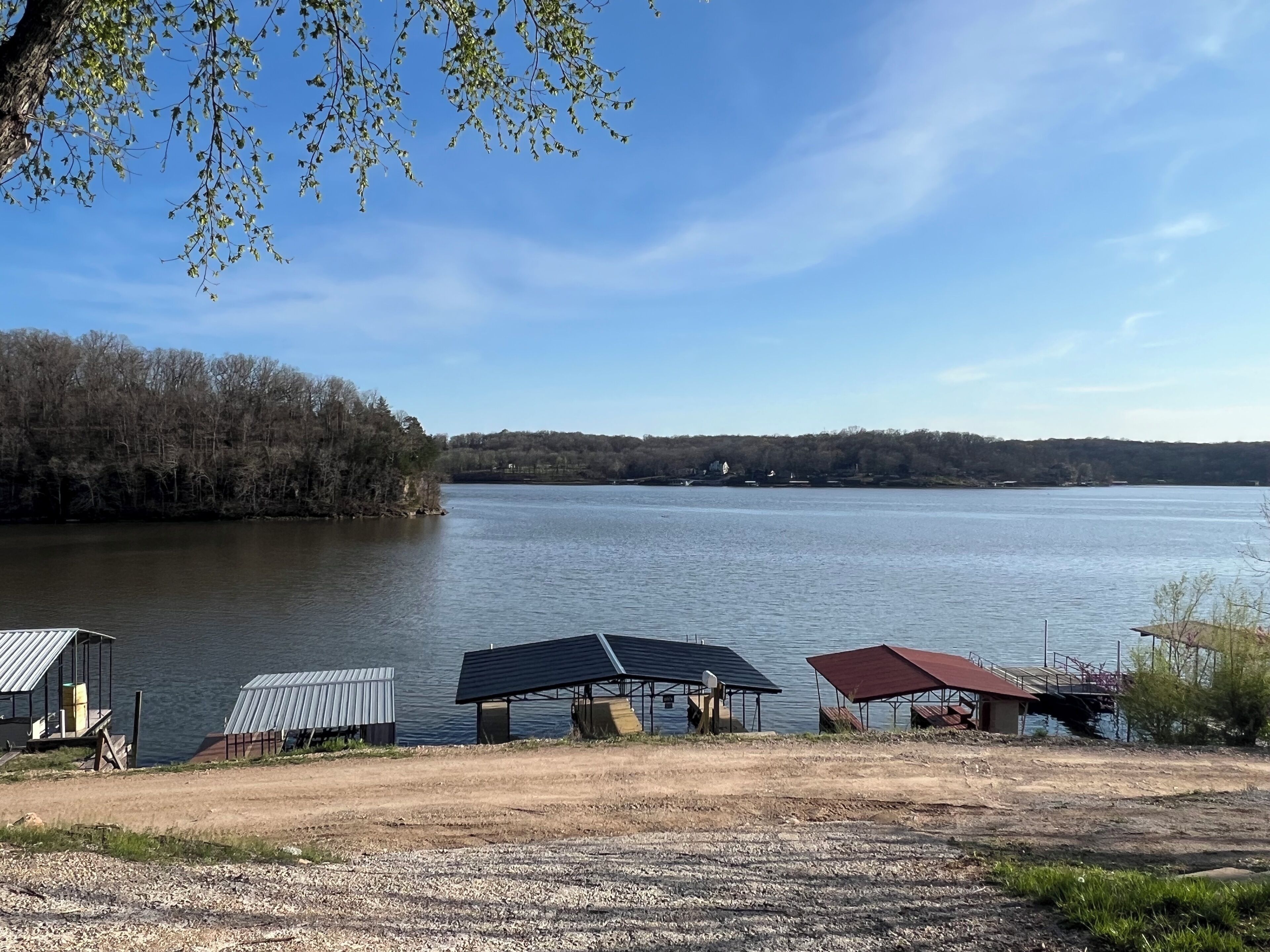 Blick auf einen See mit Kiesfläche, einem überdachten Pavillon und einem kleinen Schuppen.