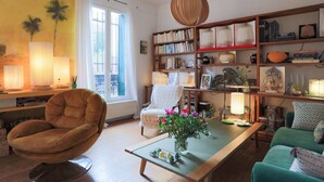 Un salon chaleureux avec un fauteuil confortable, une table basse en bois ornée d’un vase de fleurs, une bibliothèque remplie de livres et d’objets décoratifs, et une grande fenêtre avec des rideaux.