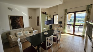 A cozy living room with a sofa, a dining table, and chairs. There is a kitchen area with cabinets and a stove. A large window offers a view of the outdoors. - House for 12 with Pool (Masone Pardu)