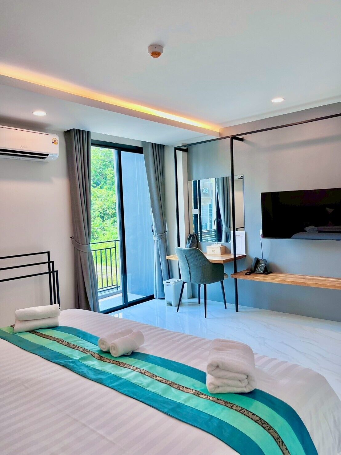 Habitación de hotel moderna con una cama grande, un escritorio, una silla, un televisor y un balcón con vistas a zonas verdes.