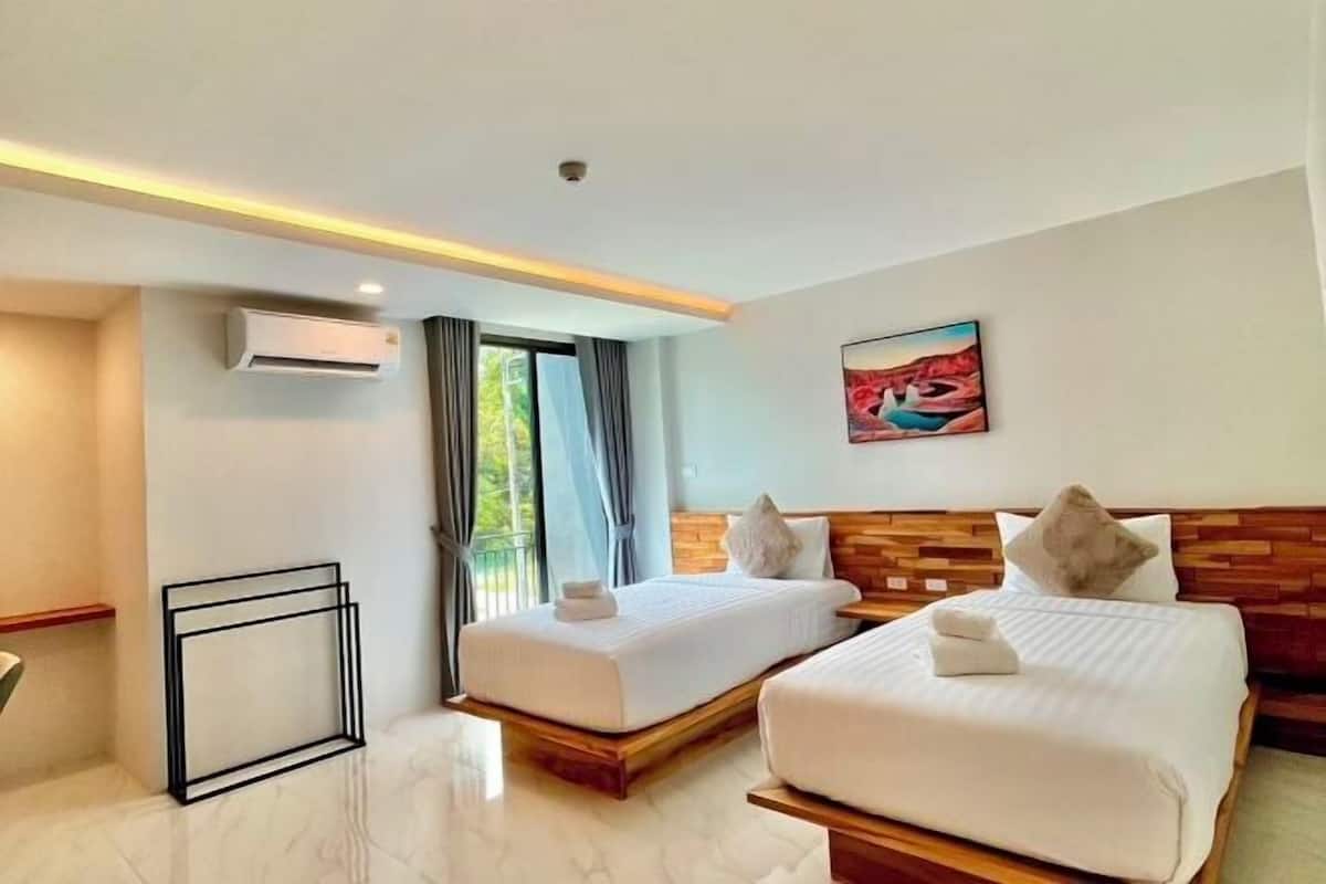 A hotel room with two beds, a painting, an air conditioning unit, and a view of greenery outside.