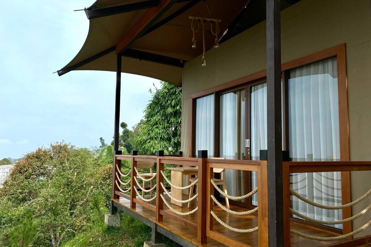 Un balcon en bois avec des balustrades en corde, de grandes fenêtres avec des rideaux et une vue sur la verdure et les arbres.
