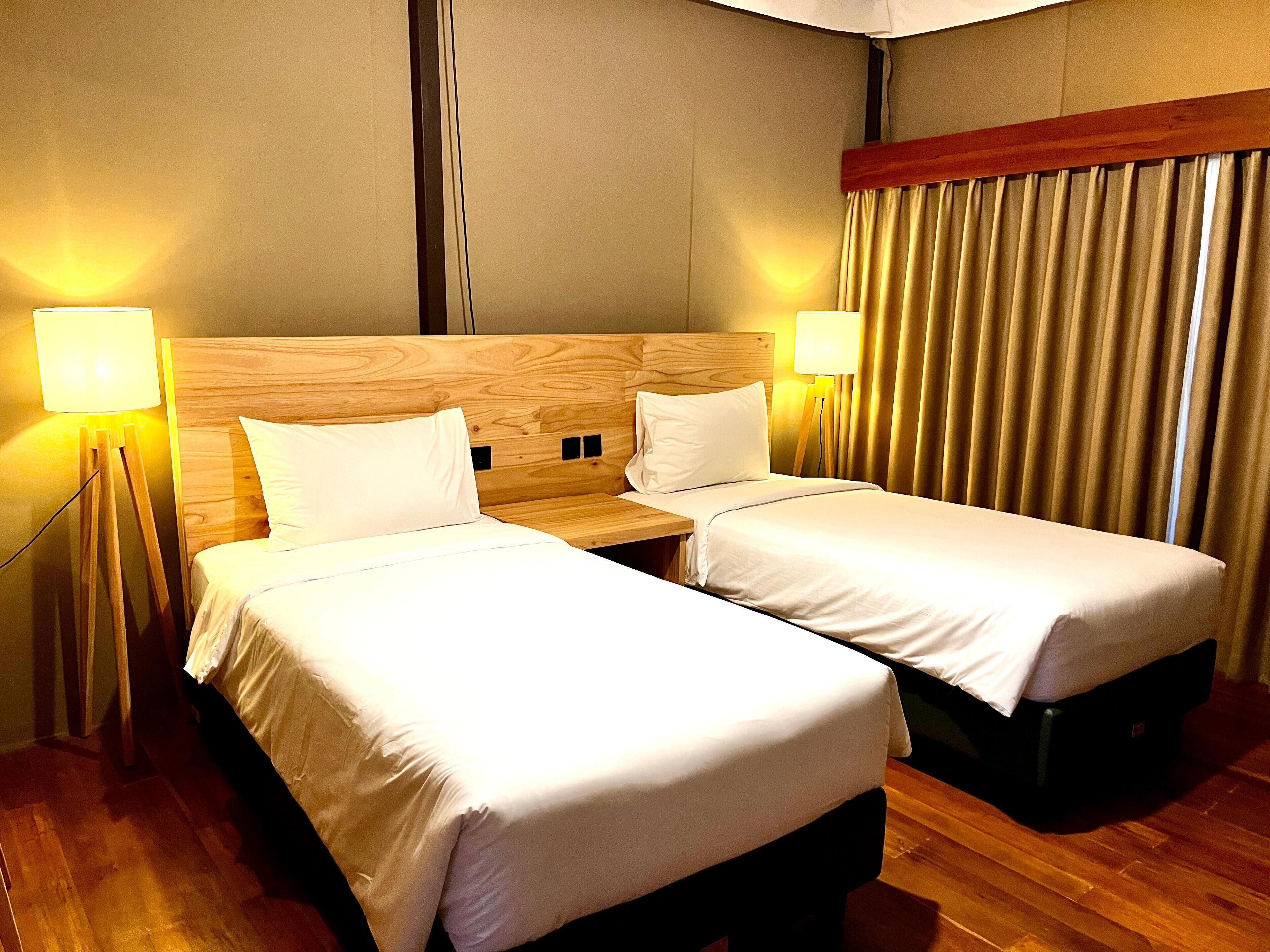 Una habitación de hotel con dos camas individuales, cabecero de madera, lámparas de noche y una cortina.