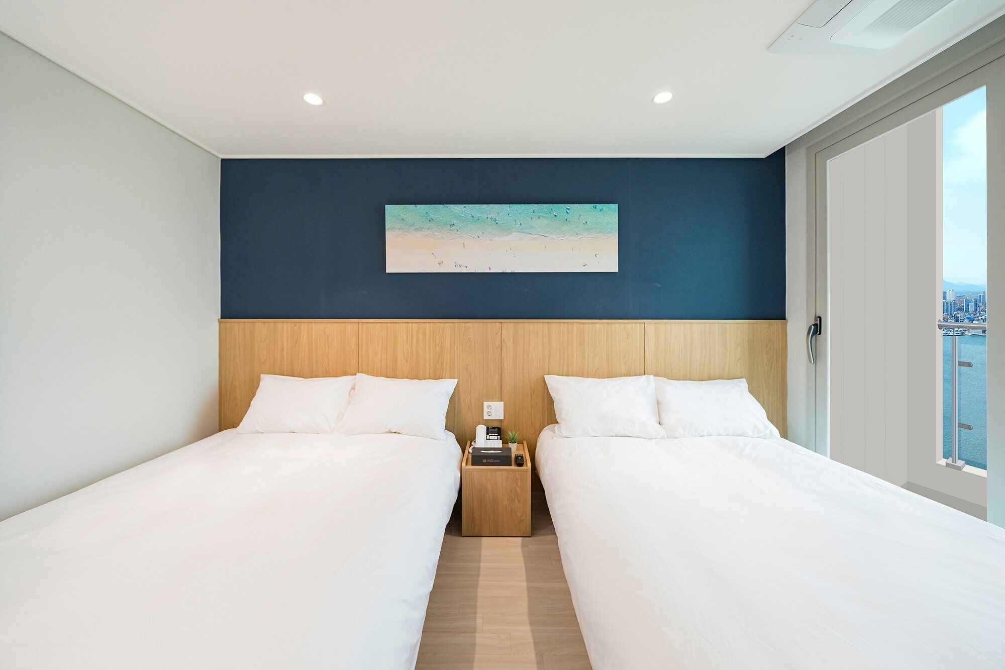 A hotel room with two beds, a nightstand, a painting of a beach, and a city view through a window.