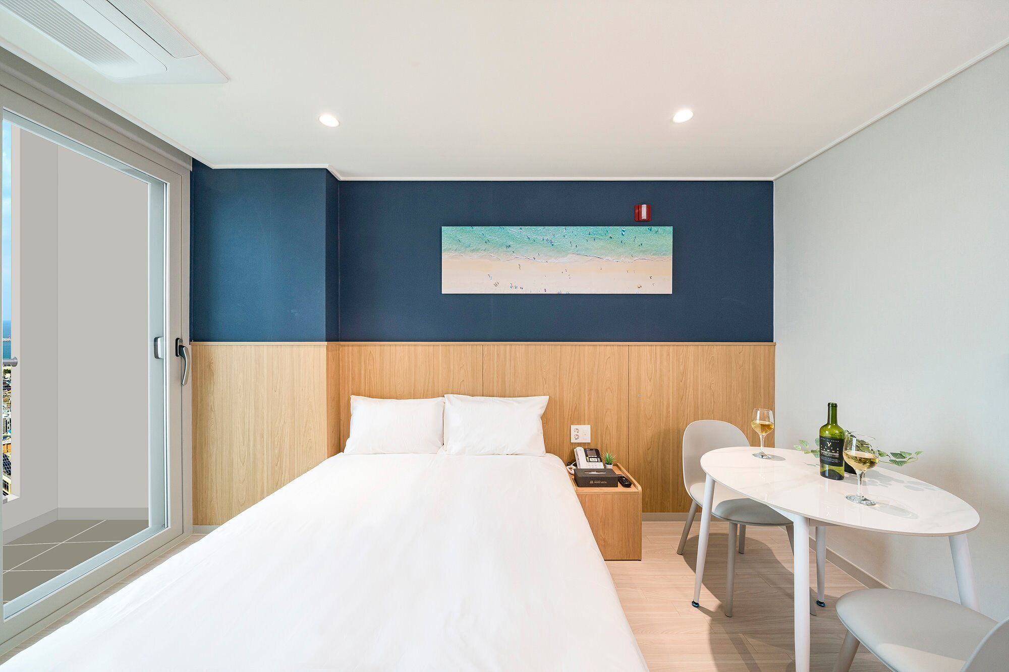 A hotel room with a bed, a small table with a chair, a painting of a beach, and a window with a view.