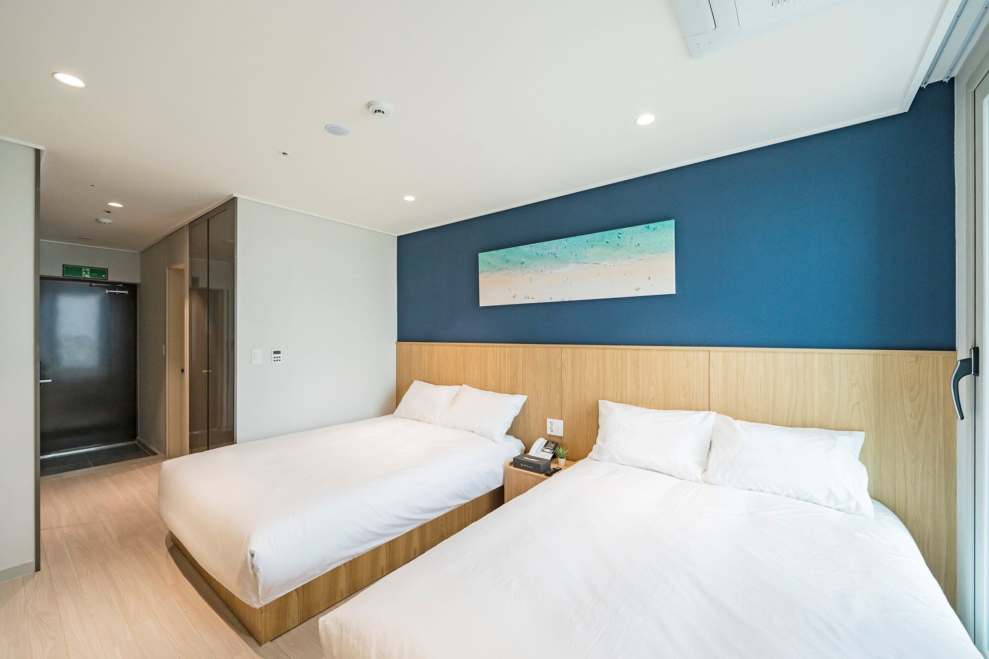 Habitación de hotel con dos camas, un cabecero de madera, un cuadro de una playa y una mesita de noche con un teléfono y una planta.