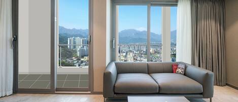 A modern living room with a grey sofa, a coffee table with books, and a view of a cityscape through large windows.