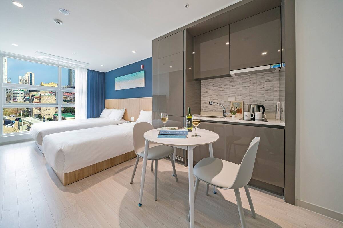 A hotel room with two beds, a dining table with chairs, a kitchen area, and a large window offering a city view.
