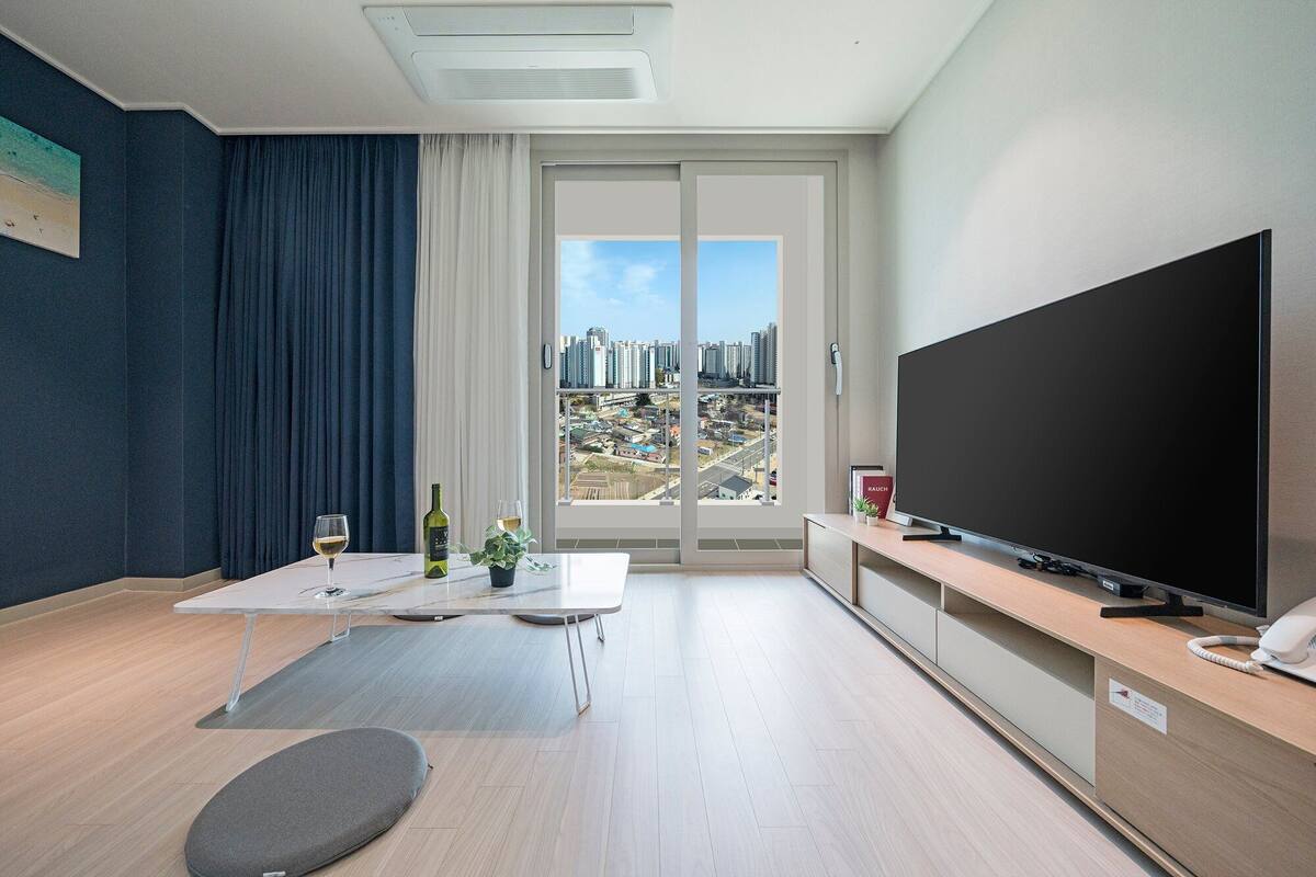 A modern living room with a large flat-screen TV, a dining table with two wine glasses, a sofa, and a view of a cityscape through a large window.