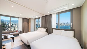 Habitación de hotel con dos camas, un sofá y grandes ventanales con vistas a la ciudad.