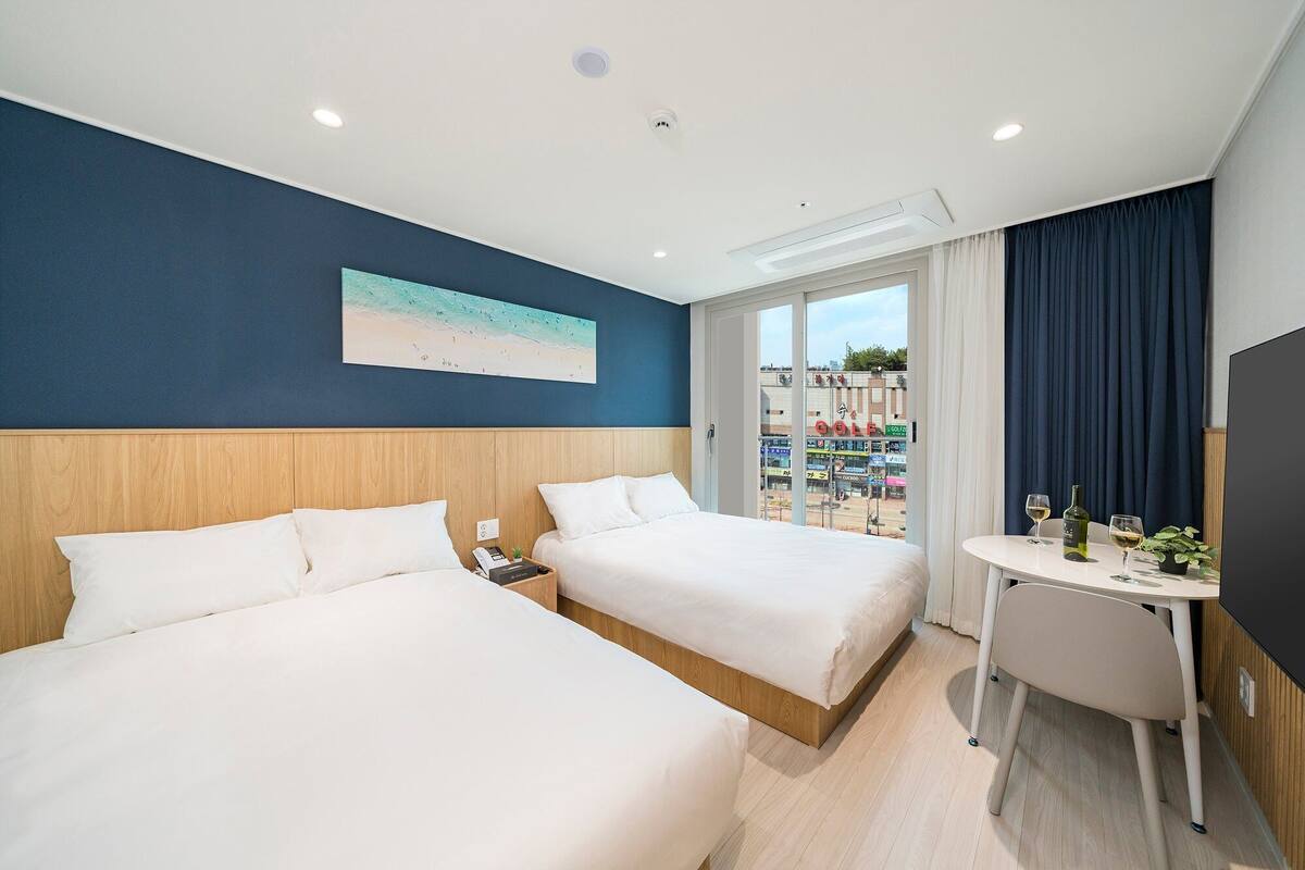 A hotel room with two beds, a desk with a chair, a painting on the wall, and a view of the outside.