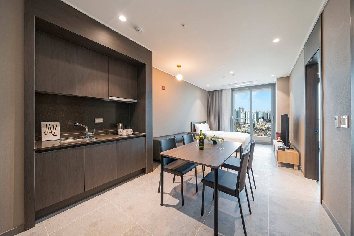 A modern hotel room with a dining area, a kitchen, and a large window offering a city view.