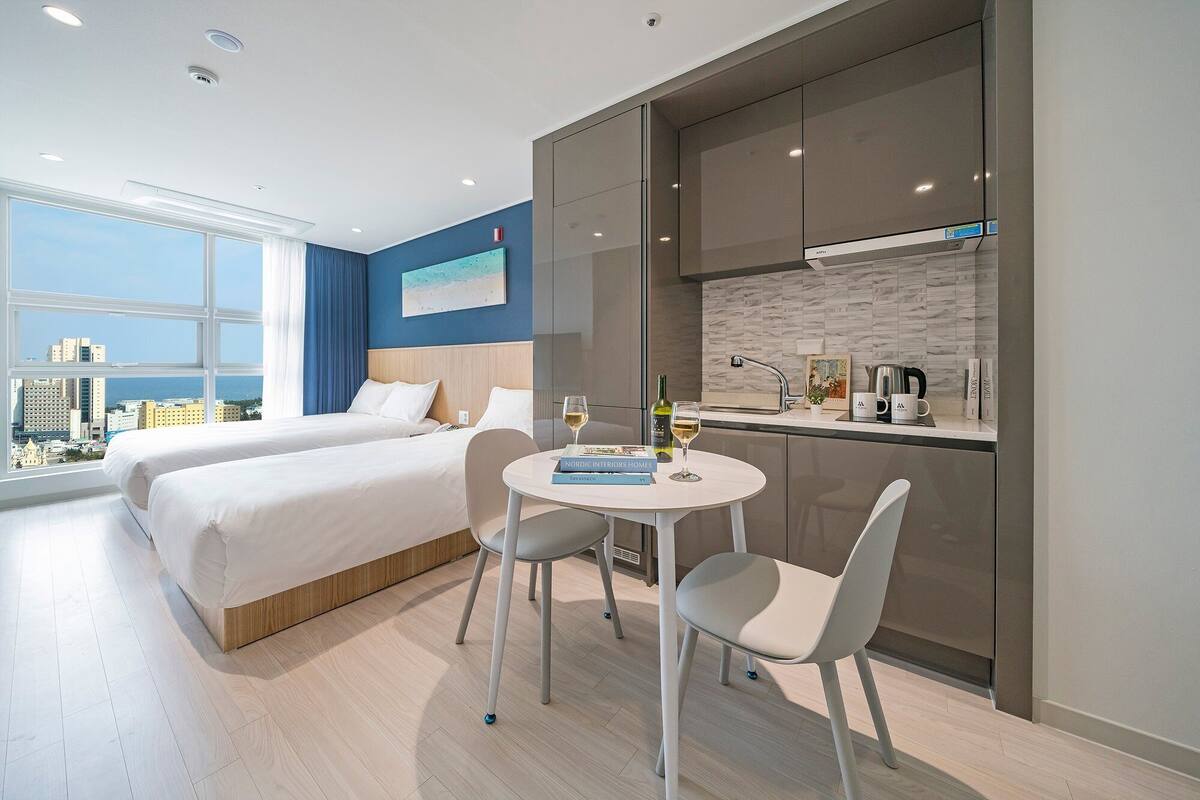 A modern hotel room with two beds, a dining area, and a kitchen.