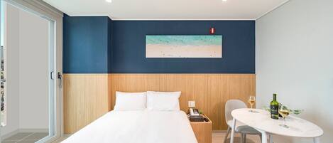 Habitación de hotel con cama, mesita de noche con teléfono y botella, una silla y una pequeña mesa con una copa de vino.