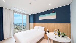 Habitación de hotel con una cama, una mesita con una botella y un vaso, un cuadro de una playa y vistas a un paisaje urbano.