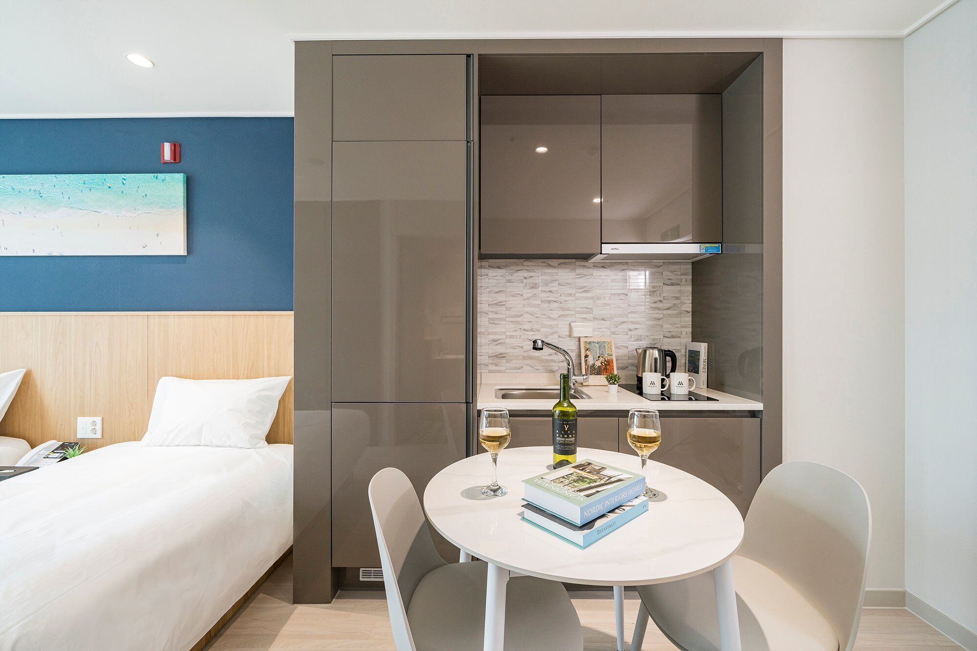 Una habitación de hotel moderna con una cama, una kitchenette pequeña, una mesa redonda con dos sillas y un cuadro en la pared.