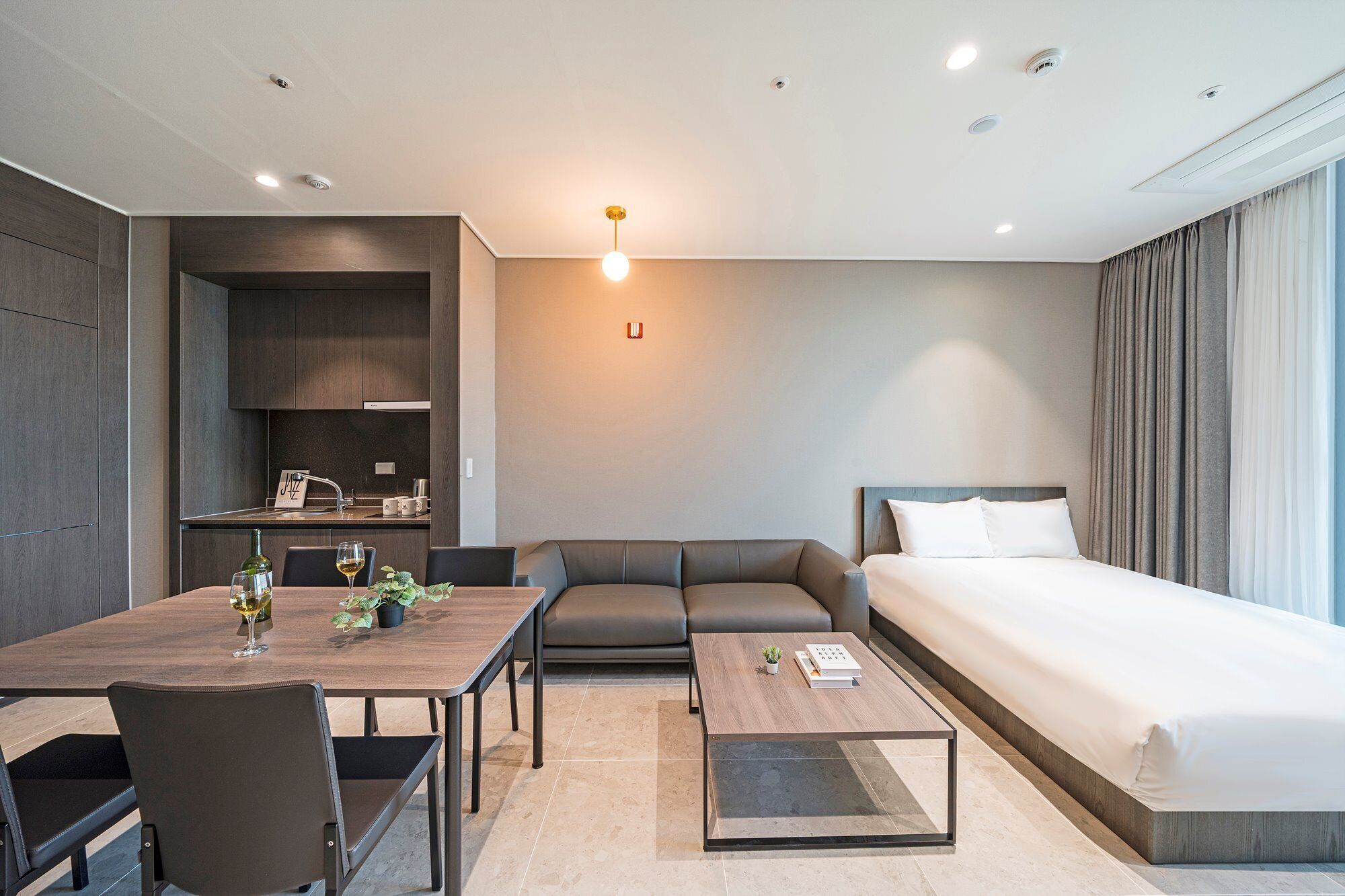 A modern hotel room with a bed, a sofa, a dining table with chairs, and a small kitchen area.