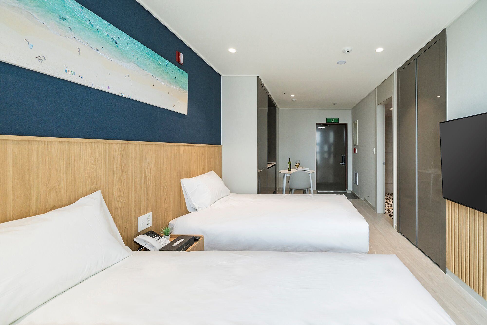 Habitación de hotel con dos camas, un cuadro grande de una playa, una mesita con una botella y un vaso, y un televisor en la pared.