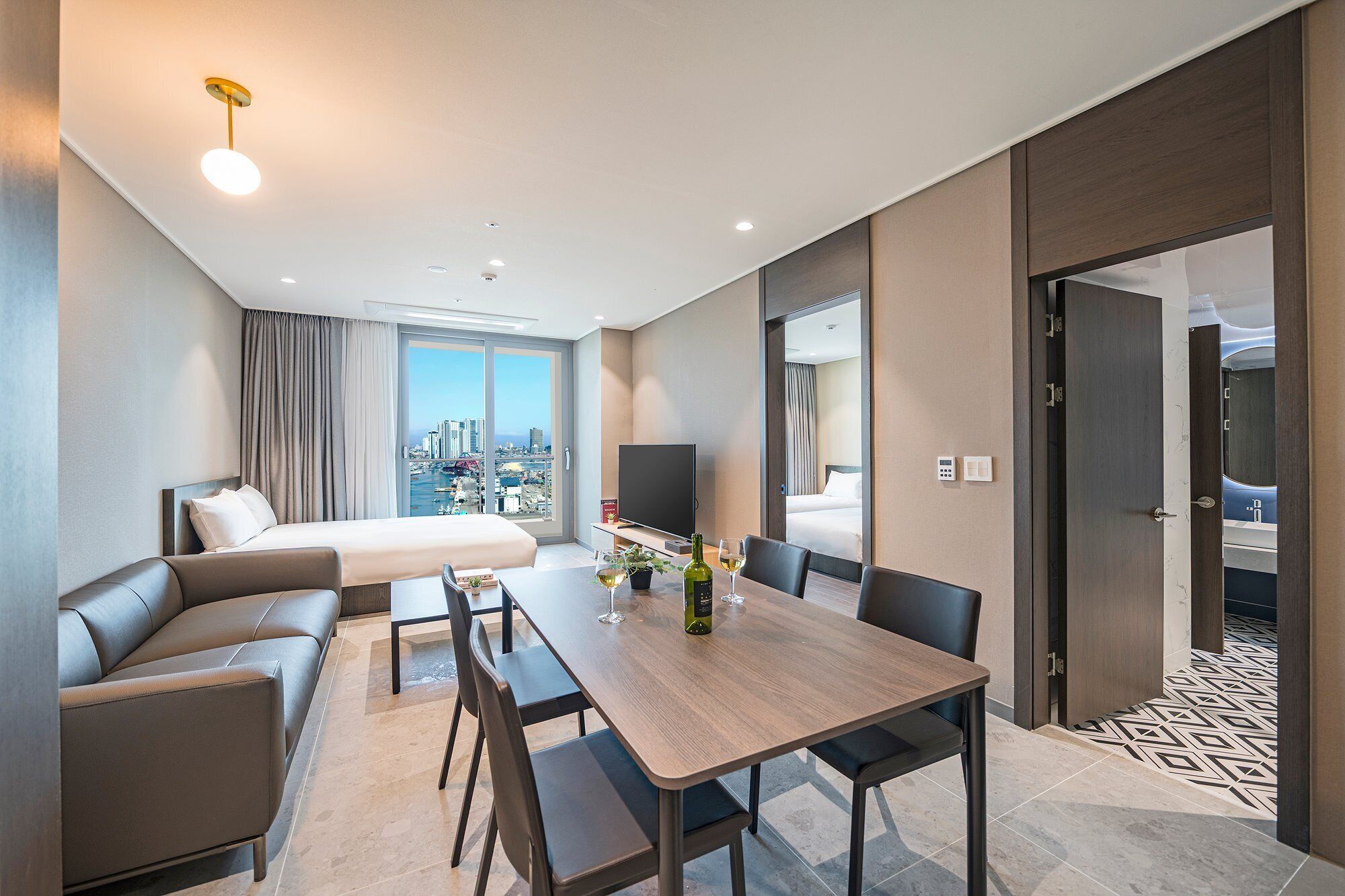 Una habitación de hotel moderna con cama, sofá, mesa de comedor con sillas, televisor y vista a la ciudad.