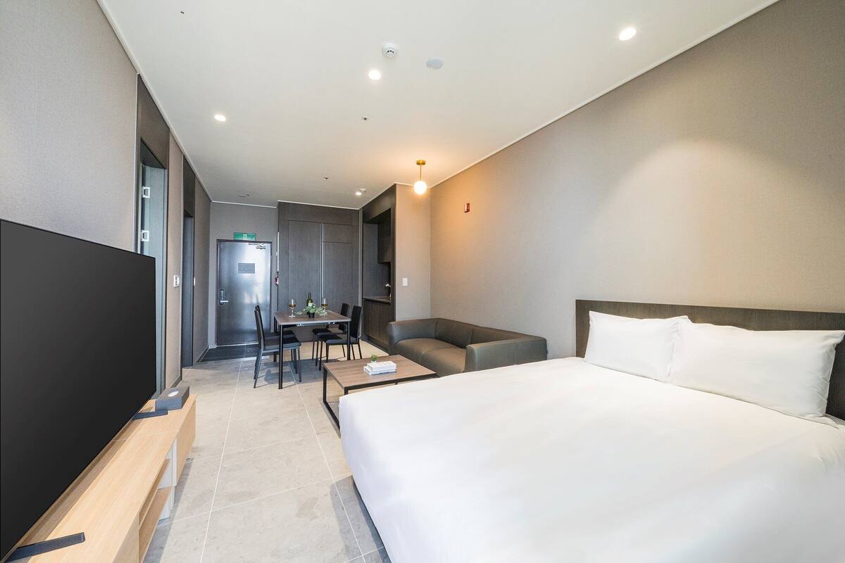 A modern hotel room with a large bed, a desk with a chair, a sofa, and a TV.