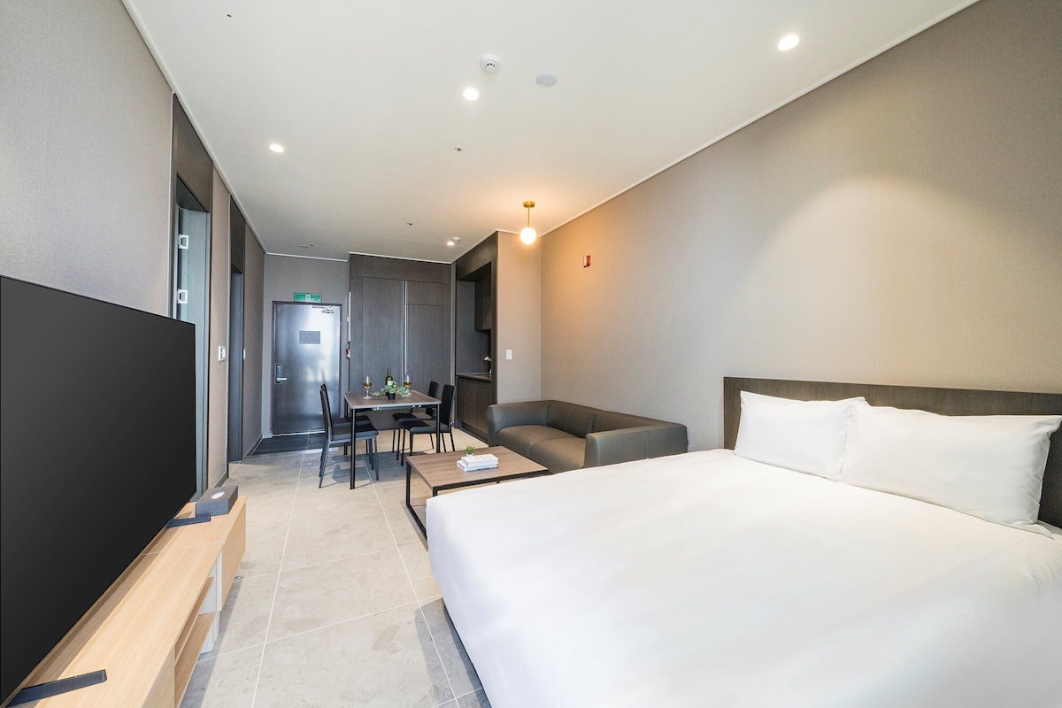 A modern hotel room with a large bed, a desk with a chair, a sofa, and a TV.