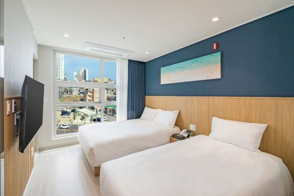 A hotel room with two beds, a large window with a city view, a flat-screen TV, and a painting on the wall.