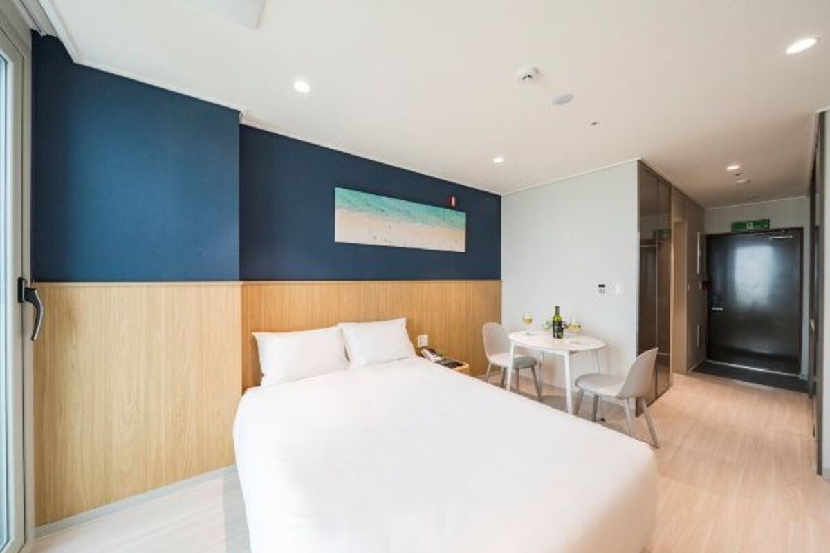 A hotel room with a large bed, a small table with chairs, a painting on the wall, and a door to the right.