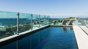 Une piscine à débordement située sur le toit, avec des garde-corps en verre, offrant une vue imprenable sur le paysage urbain et l’océan.