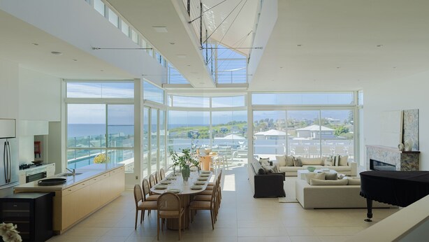 Un vaste espace de vie comprenant une salle à manger avec une table pour six personnes, un canapé et une cheminée. De grandes fenêtres offrent une vue sur la mer et le paysage environnant.