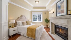 A bedroom with a bed, nightstands, a fireplace, a window, and a potted plant.