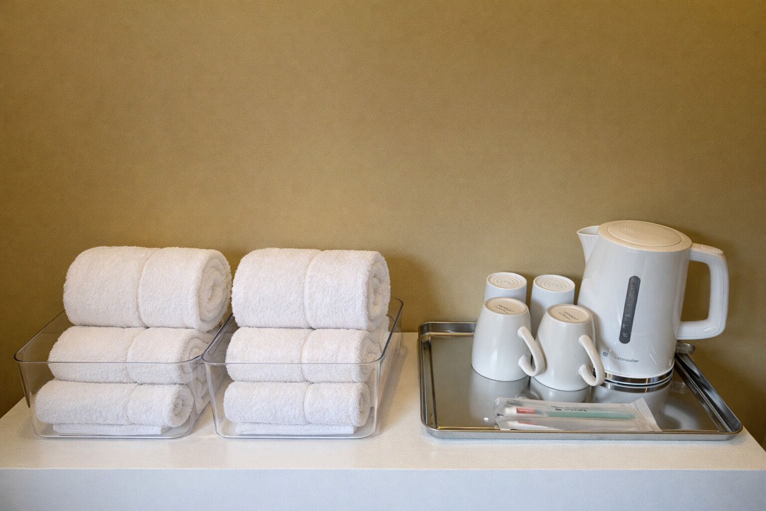 A hotel room's amenities area with neatly stacked white towels in clear containers, a tray with white mugs and a kettle, and a small tray with toiletries.